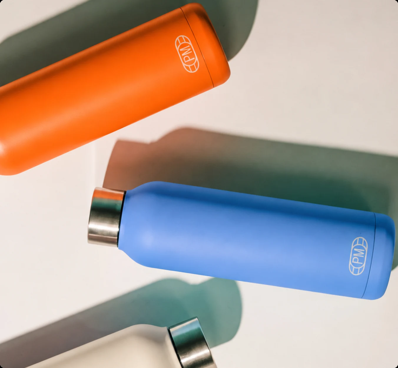 Three PropMedia water bottles — orange, blue and green, each with the PM monogram — on a white ground
